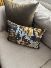 Load image into Gallery viewer, A rectangular lumbar throw pillow with a tropical floral and palm leaf pattern in shades of blue, gold, and dark brown, styled on a light gray fabric sofa next to a solid olive green pillow.