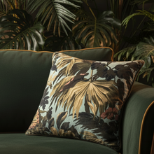 Load image into Gallery viewer, A square decorative throw pillow with a tropical palm leaf pattern in beige, brown, and muted red on a light blue background, resting on a dark green velvet armchair with gold piping, set against a backdrop of large green houseplants.
