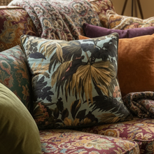 Load image into Gallery viewer, A square decorative throw pillow with a tropical palm leaf pattern in muted blue, olive, and brown tones, styled on a vintage floral sofa alongside other textured cushions.
